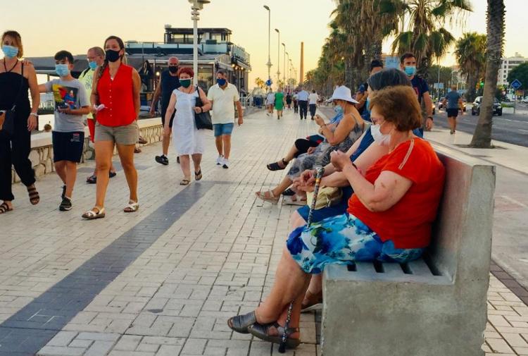 Mujeres sentadas, mientras otras personas pasean cerca de la playa, todas y todos con mascarilla.