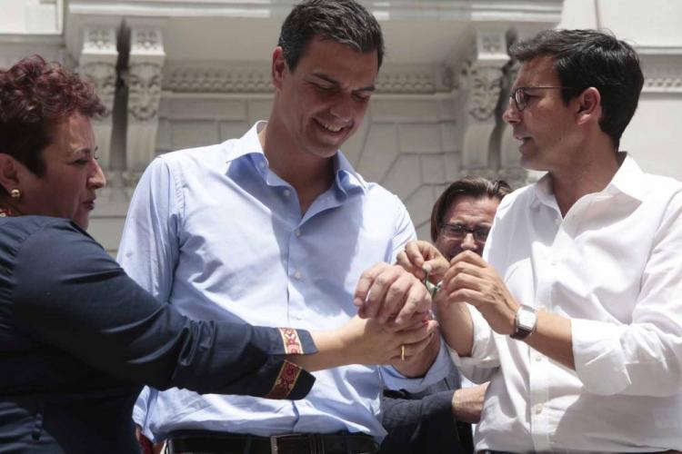 Cuenca, ayudado por Teresa Jiménez, coloca una pulsera de la candidatura socialista a Pedro Sánchez.